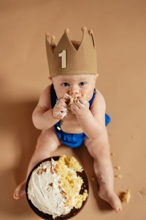 Baby wearing a gold crown with number 1 smashes first birthday cake, sitting on beige background.