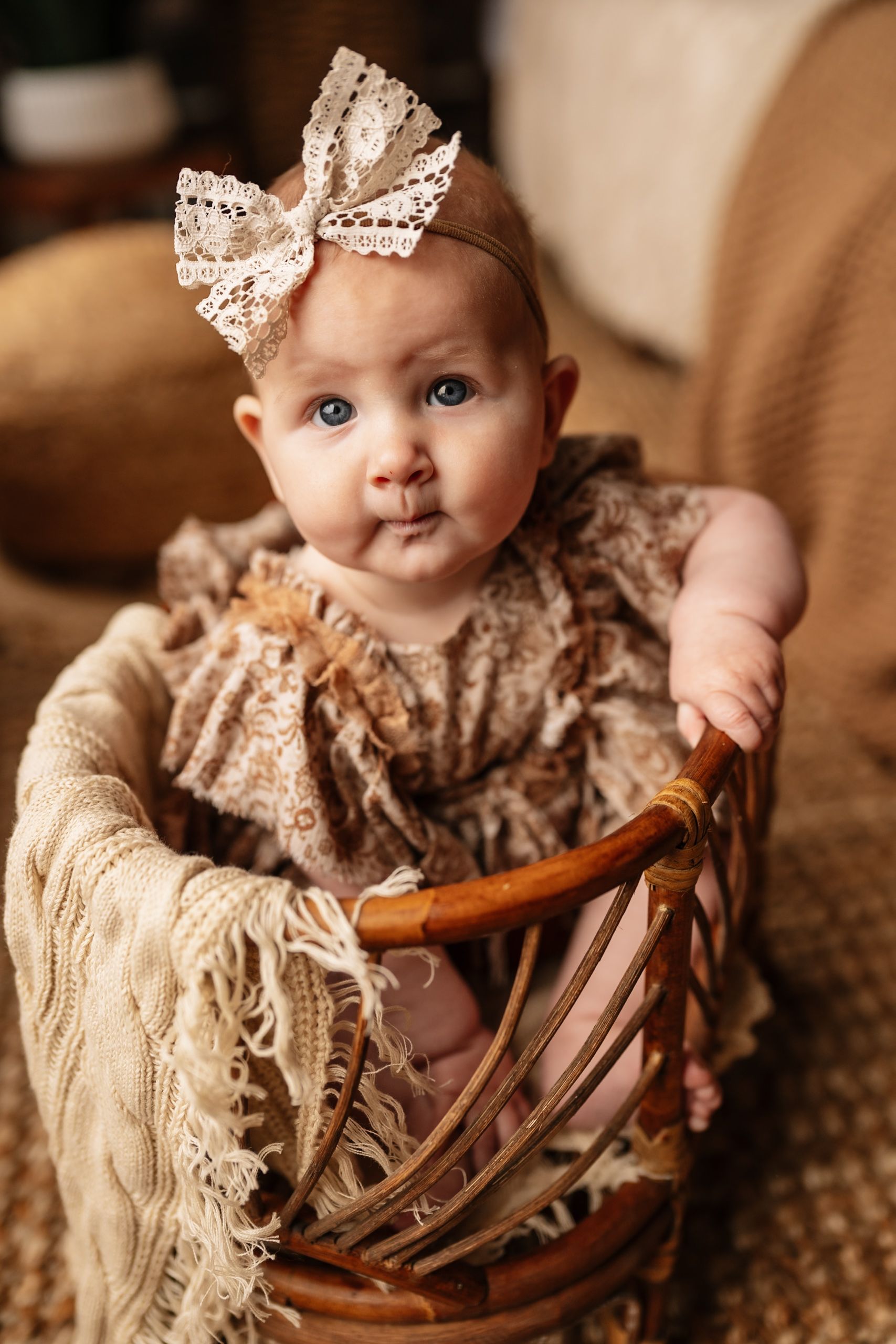 Adorable baby girl in floral dress with lace bow headband sitting in wicker basket for portrait photo.