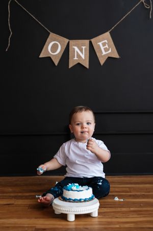 Baby boy in white shirt sitting on wood floor with blue decorated smash cake under ONE banner for first birthday.