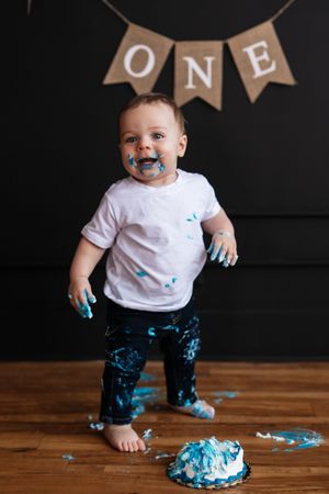 Adorable one year old boy covered in blue frosting standing next to smashed cake during first birthday cake smash.