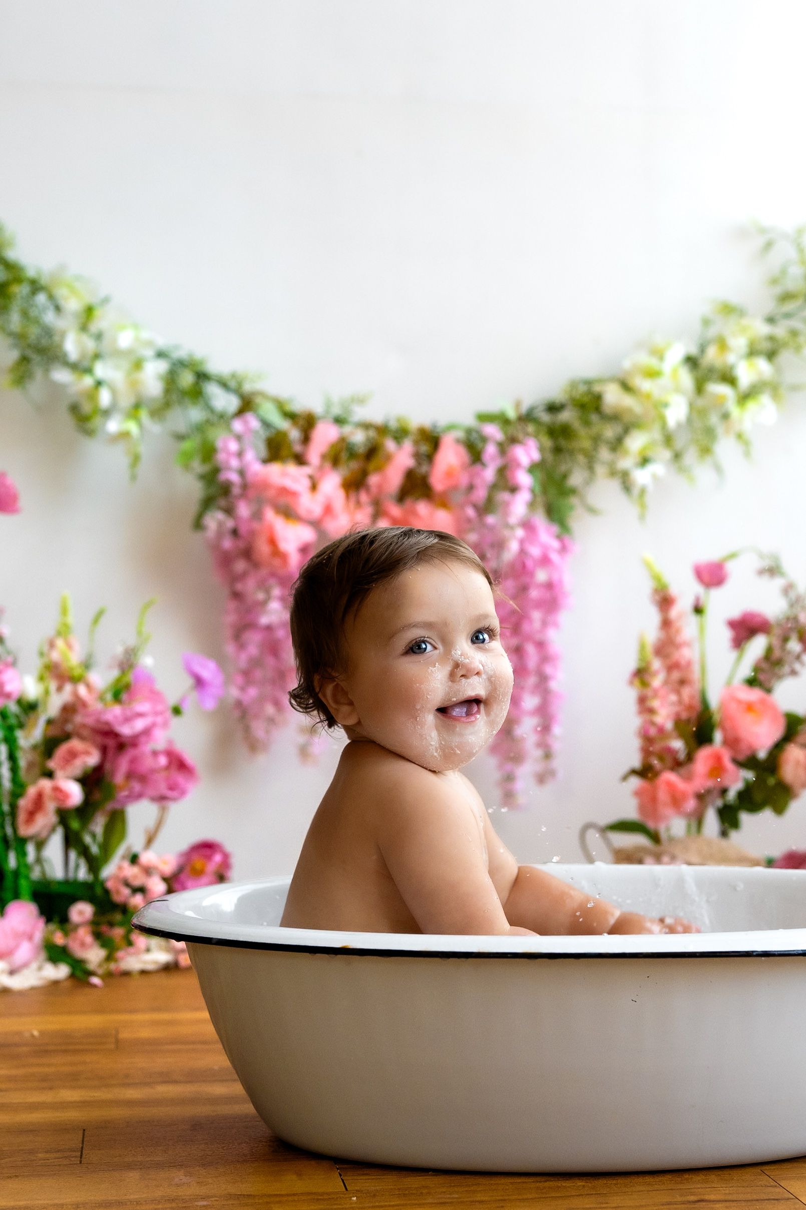 Smiling baby sitting in a white enamel tub surrounded by pink flowers and floral garland backdrop.