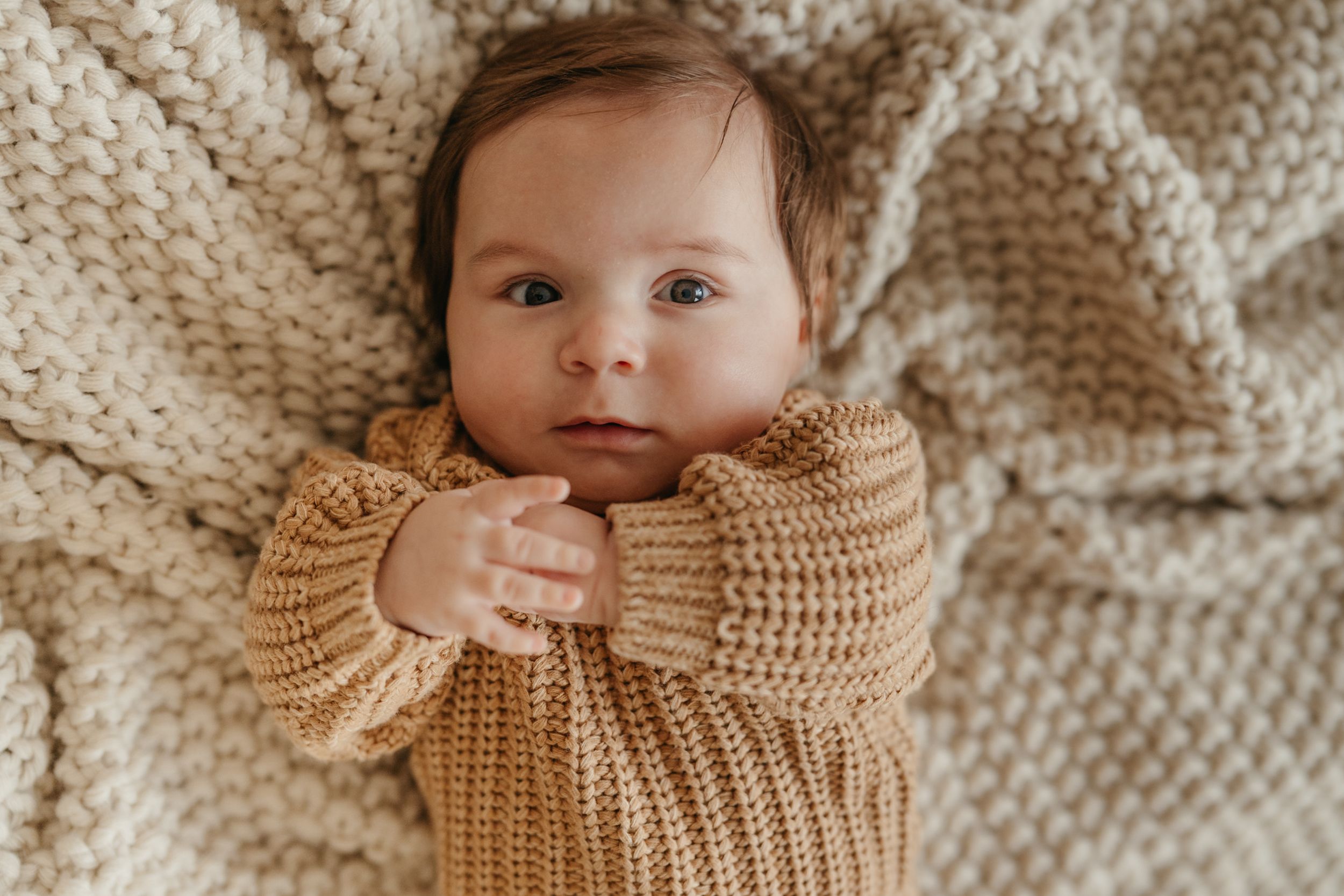 A newborn baby in a golden knit sweater lies on a cream chunky knit blanket, gazing up with bright eyes.