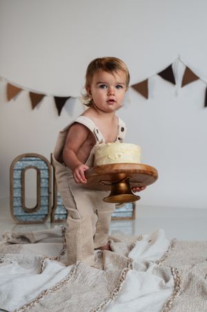 A toddler in linen overalls holds a small cake on a wooden stand at a first birthday cake smash photo session.