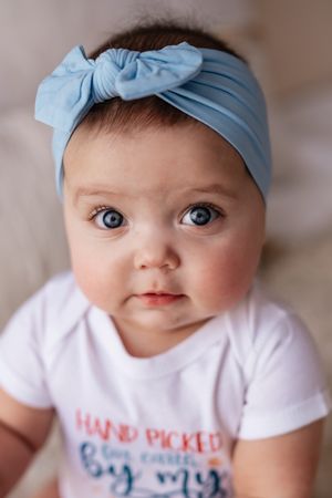 Close-up portrait of baby girl with blue eyes wearing light blue bow headband and white onesie.