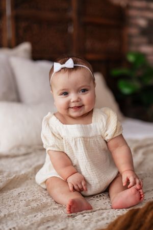 Smiling baby girl in cream romper and white bow headband sitting on a lace blanket in a cozy bedroom.