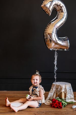 Toddler girl sitting on floor beside a large silver number 2 balloon and red roses for her 2nd birthday.