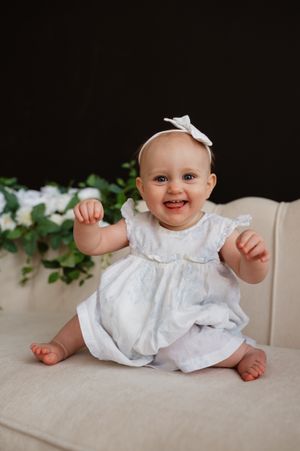 Smiling baby girl in white dress and bow headband sitting on cream sofa with dark background and greenery.