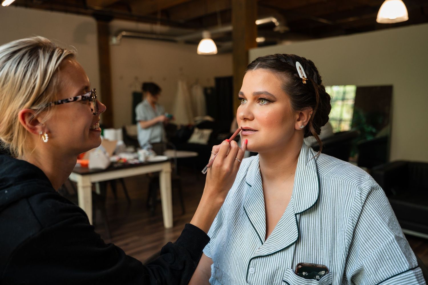 Makeup artist applying lipstick to a bride in a blue striped robe during wedding preparations.