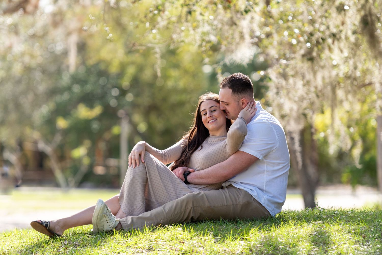 Couple sitting together on green grass in a sunny park, sharing a tender romantic moment surrounded by trees.