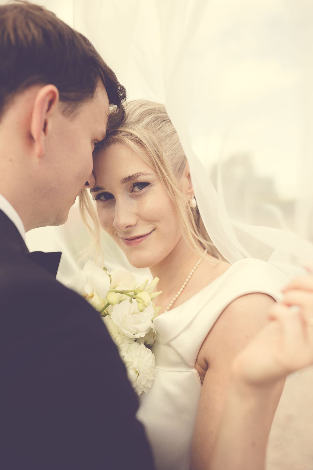 Bride and groom share an intimate moment, bride smiling softly in white dress and veil holding white bouquet.