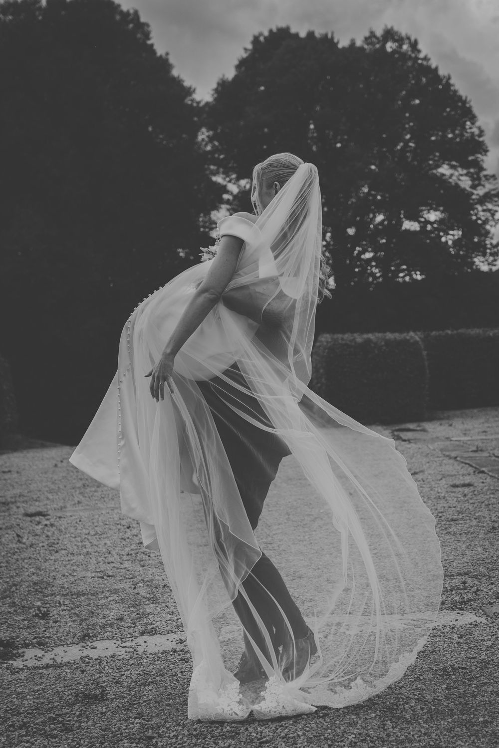Black and white photo of a bride in a flowing wedding dress and veil billowing in the wind outdoors.
