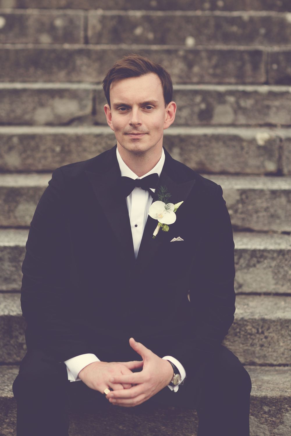 Groom in black tuxedo with white boutonniere sits on stone steps, smiling confidently at camera.