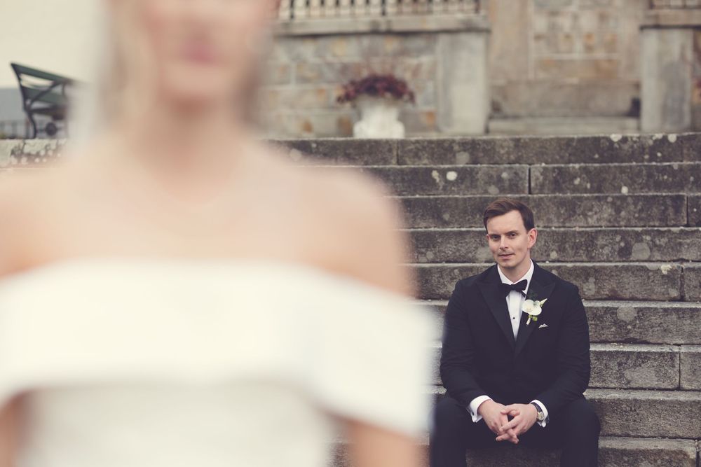 Groom in black tuxedo sits on stone steps gazing at blurred bride in white off-shoulder wedding dress.
