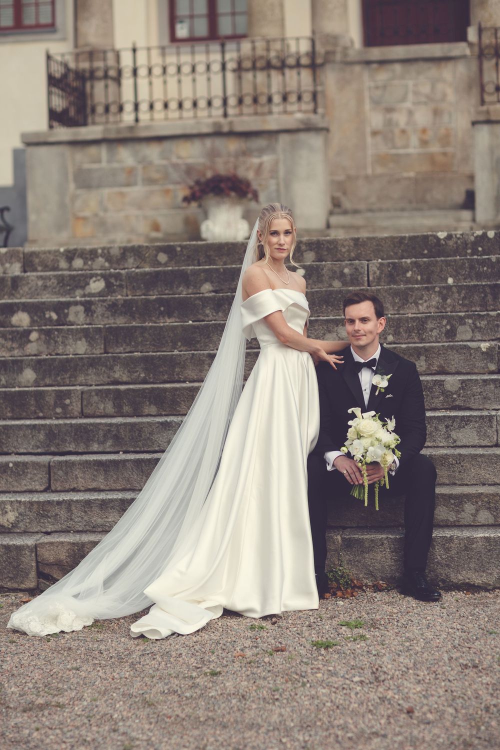 Elegant bride in off-shoulder gown with veil stands beside groom in tuxedo on stone steps of historic venue.