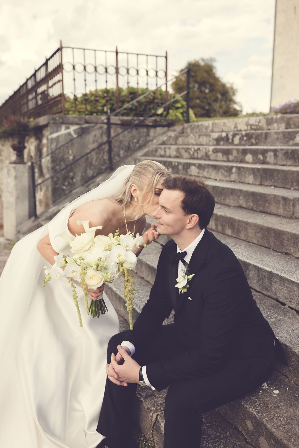 Bride and groom share a tender kiss on stone steps, bride holding white floral bouquet in elegant wedding attire.