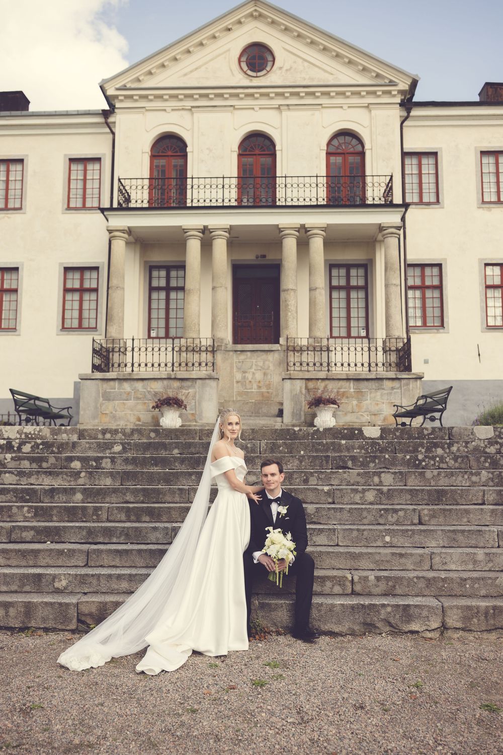 Bride and groom posing on stone steps before a grand white neoclassical mansion on their wedding day.