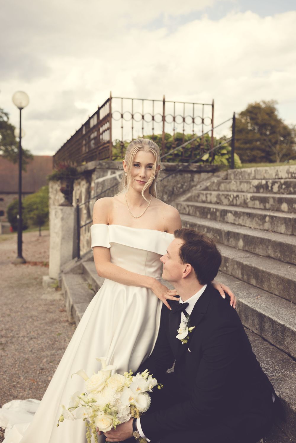 Bride in off-shoulder white gown with groom kneeling beside her holding bouquet on stone steps.