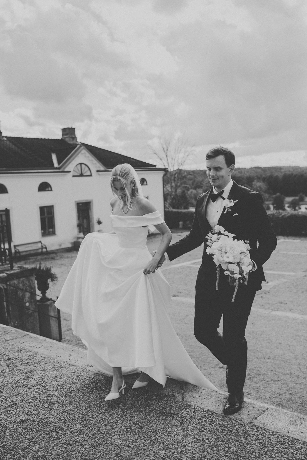 Black and white photo of bride and groom walking hand in hand outside a white estate building on their wedding day.