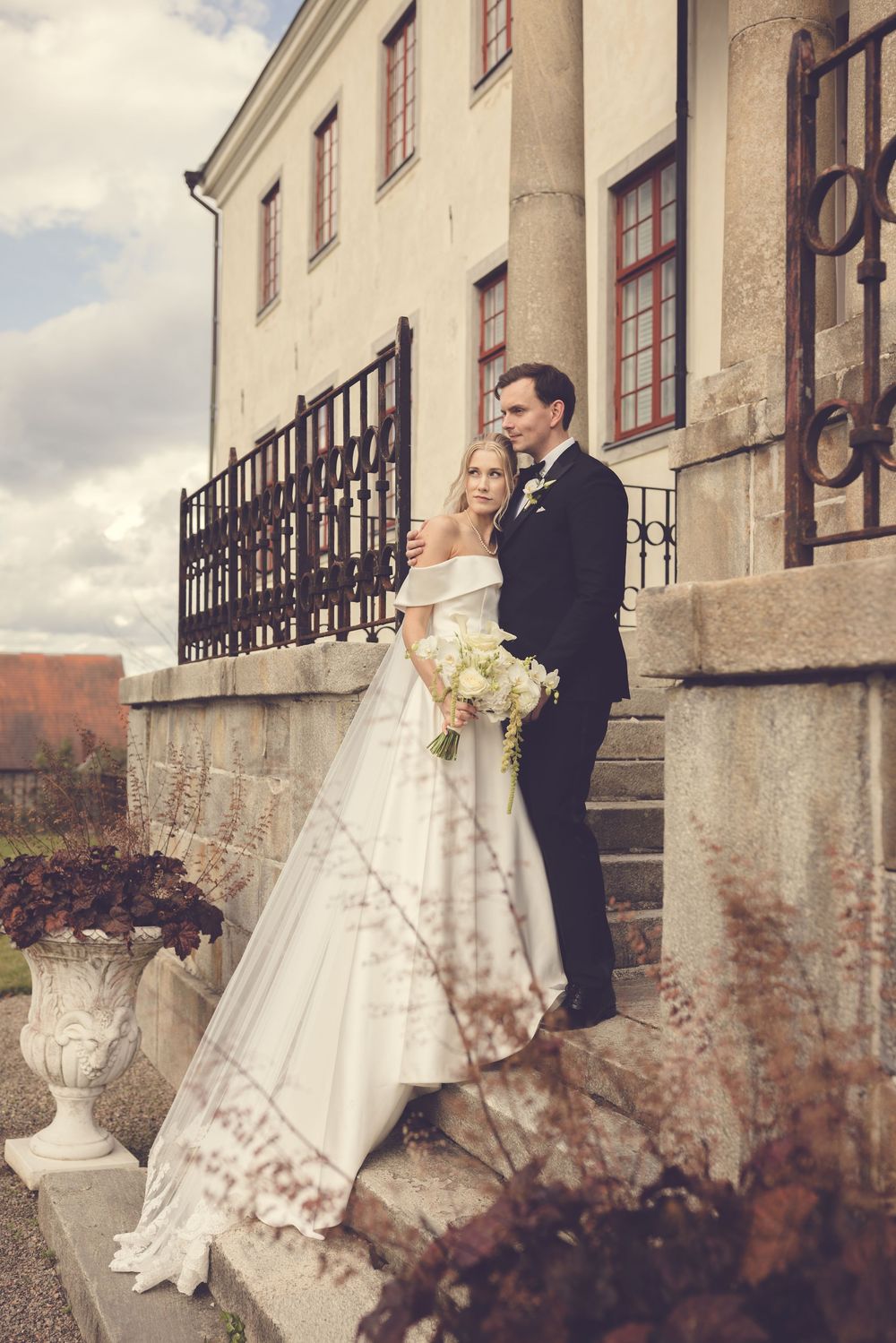 Elegant bride and groom posing on stone steps outside a historic manor, bride holding white bouquet in flowing off-shoulder gown.
