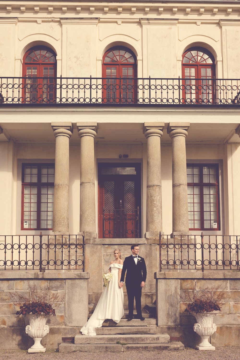 Elegant wedding couple standing at entrance of historic neoclassical manor with columns and ornate balcony.