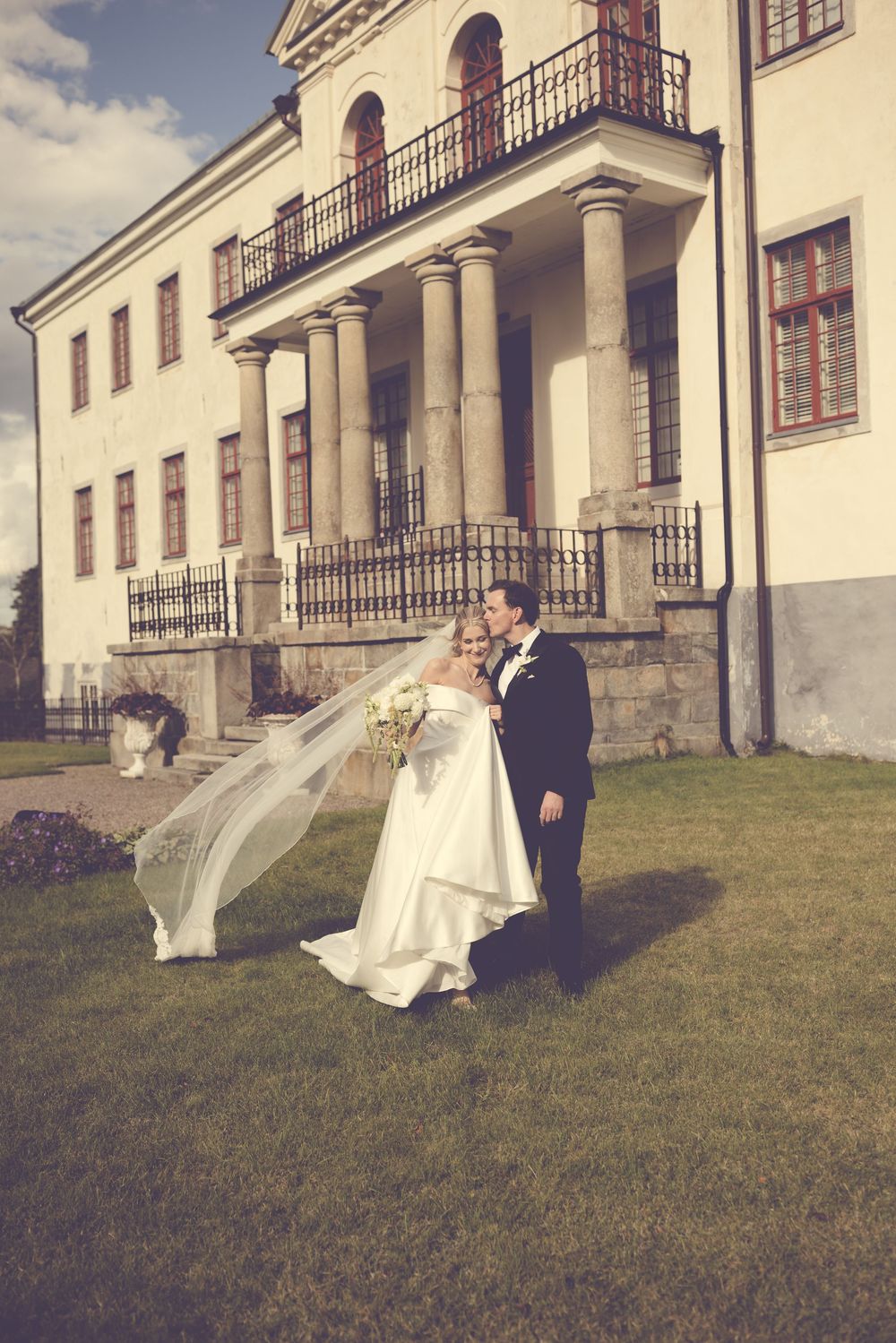 Bride and groom walking on green lawn in front of elegant white manor house with columns, veil flowing in the wind.