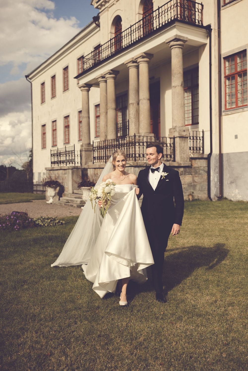 Bride and groom walk hand in hand across green lawn before a grand white neoclassical manor with tall columns.
