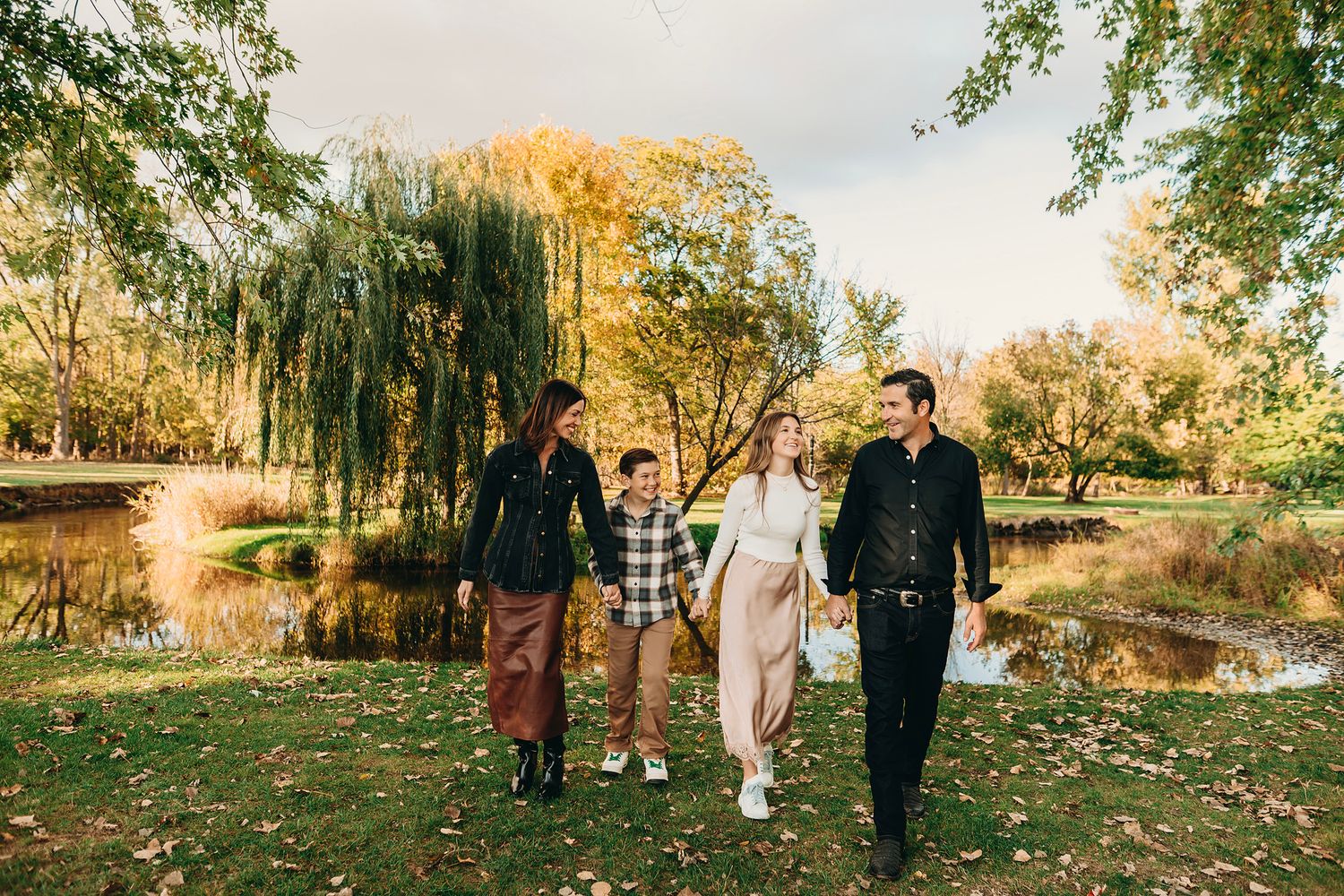 Family of four walking hand-in-hand in an autumn park with colorful trees and a serene pond in the background.