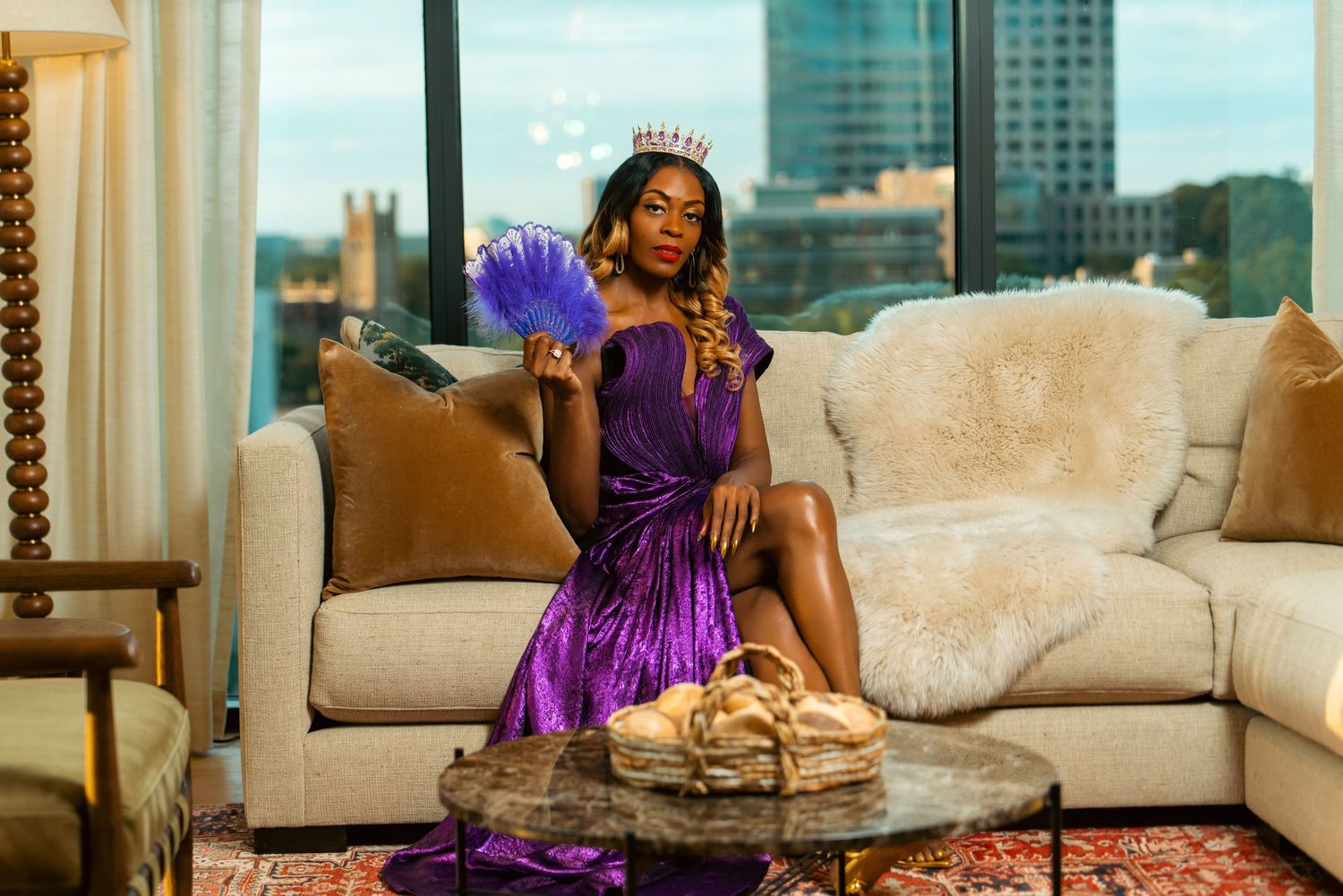 A woman in a purple sequin dress and tiara holds a feather fan while seated on a beige sofa in a luxury hotel suite.