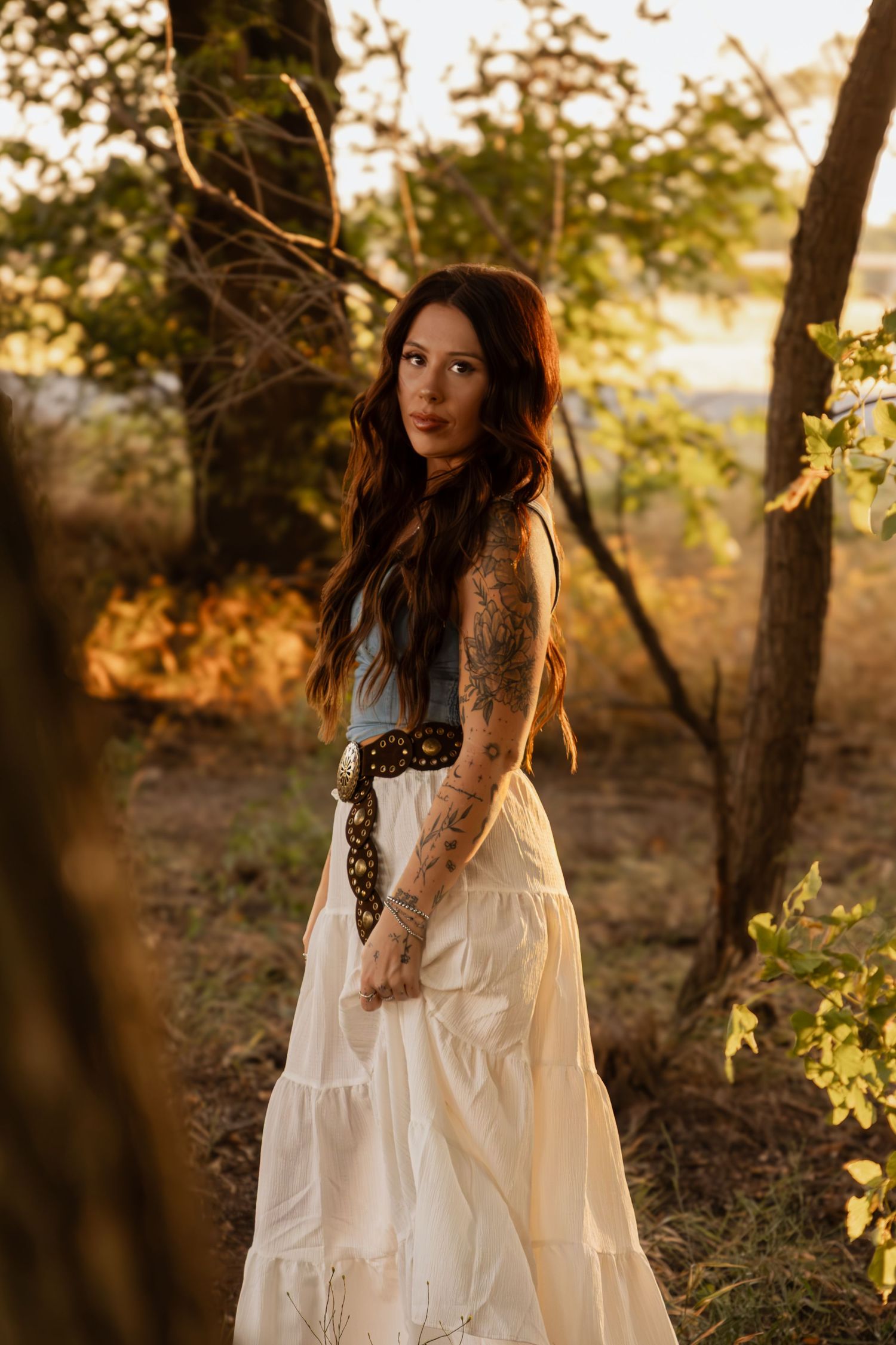 Woman in a flowing white dress posing outdoors among trees during a warm, cinematic portrait session.