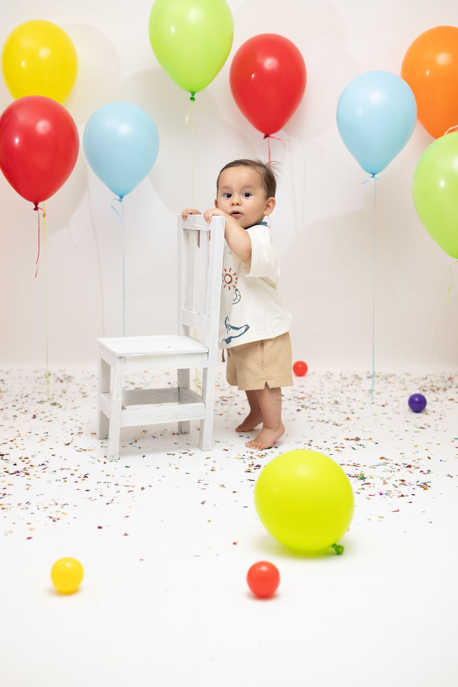 A baby boy stands holding a white chair surrounded by colorful balloons and confetti during a first birthday celebration.