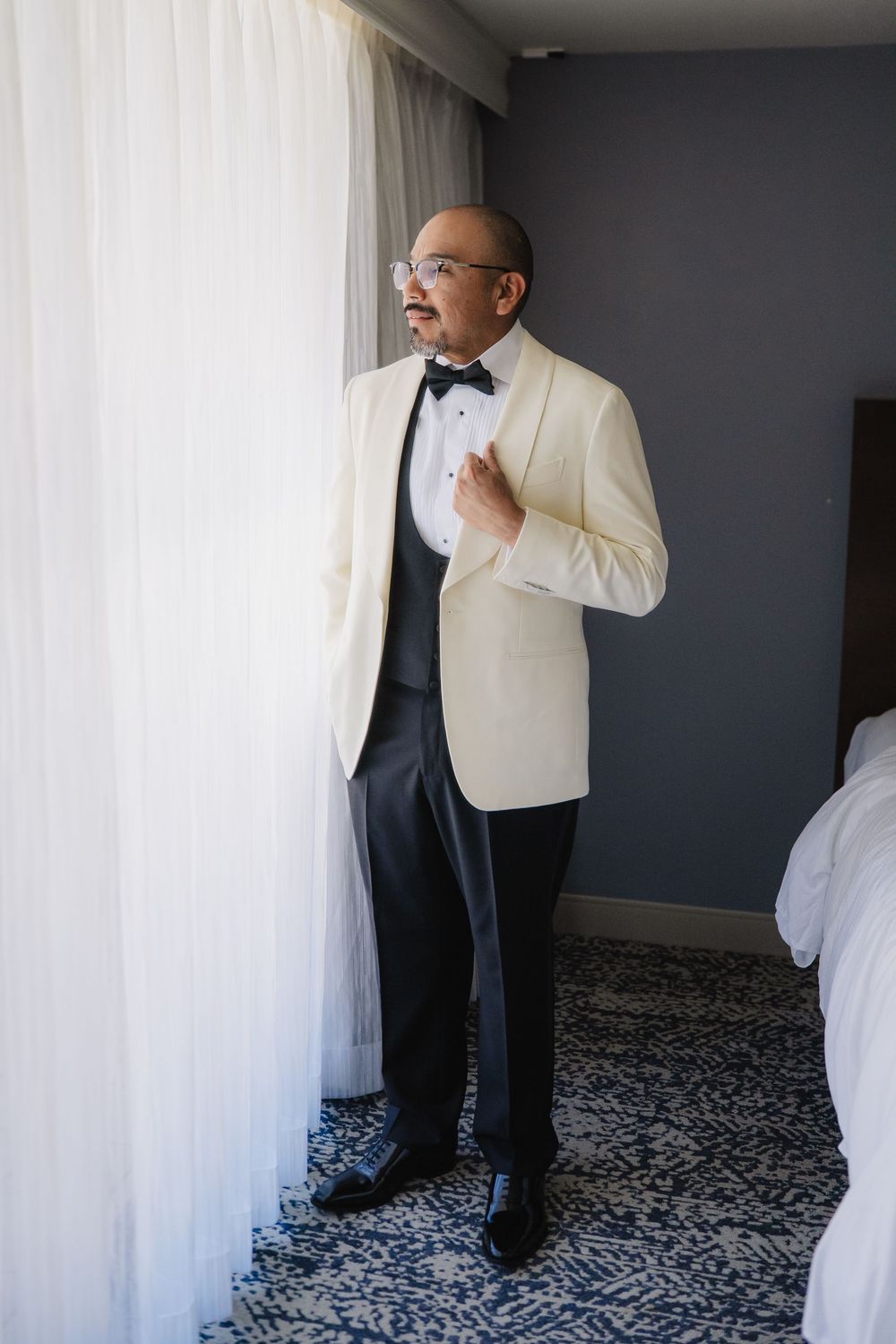 Groom in white tuxedo jacket and black bow tie adjusting lapel near curtained window in hotel room.