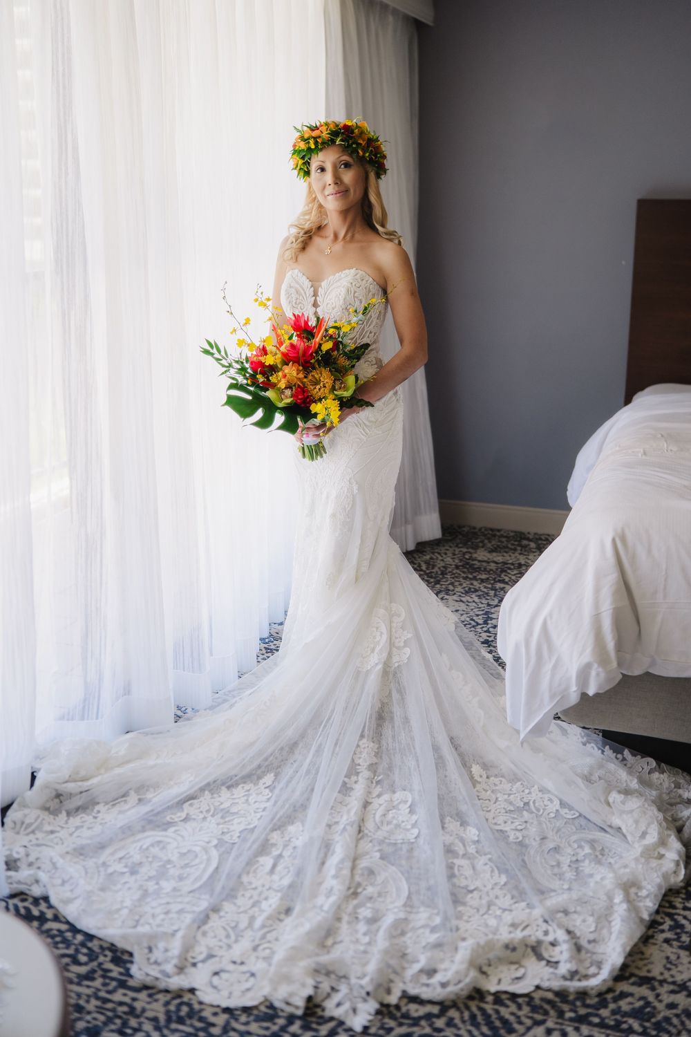 Bride in strapless lace mermaid gown with floral crown holds colorful bouquet by window.