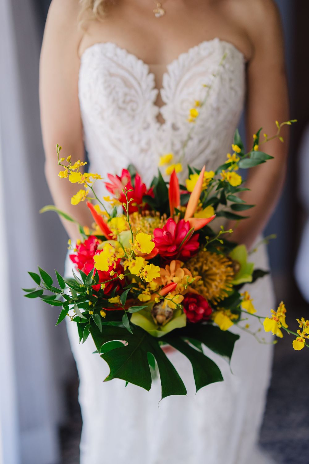 Bride in white lace gown holding vibrant tropical wedding bouquet with red, yellow, and orange flowers and green leaves.