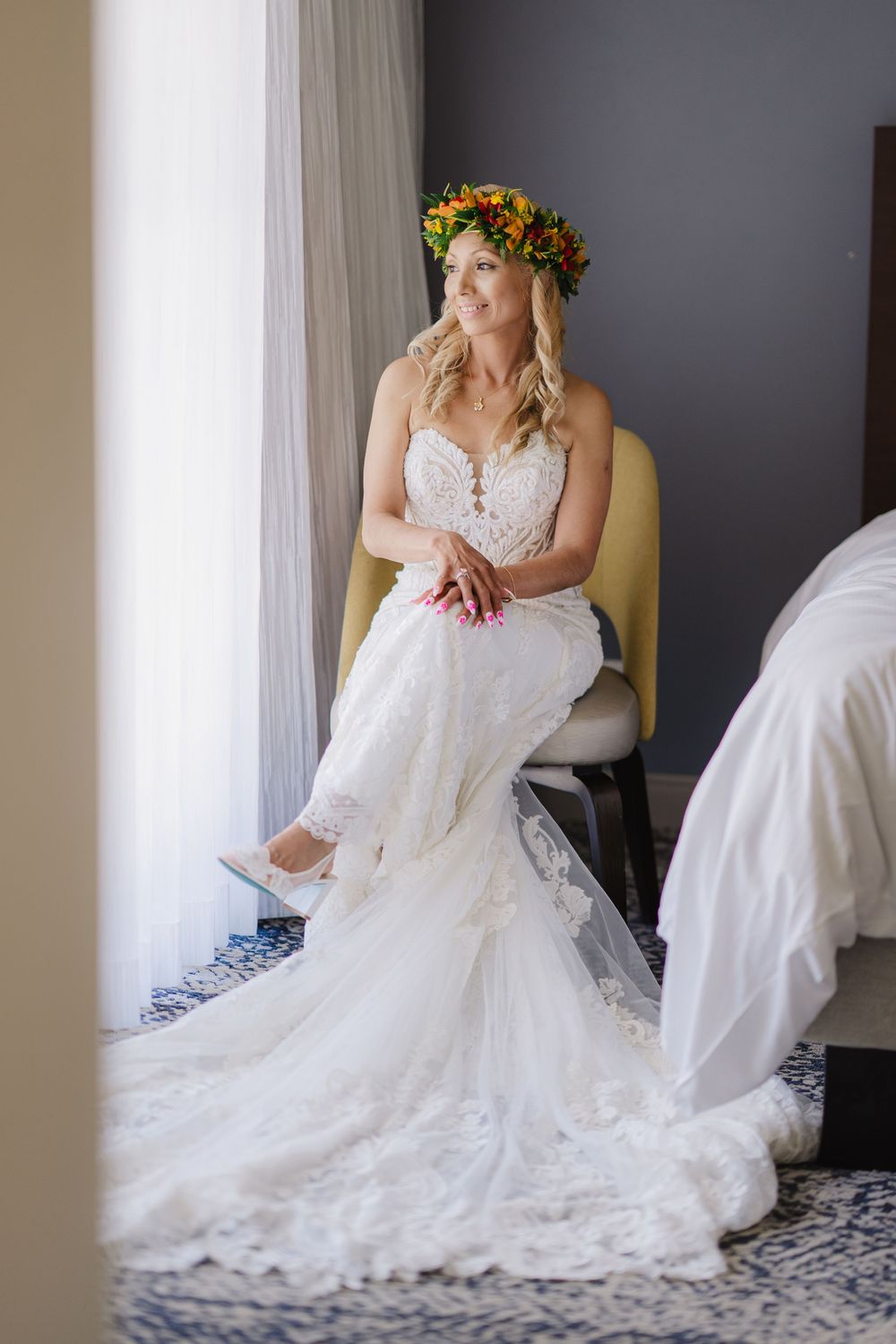 Bride in white lace wedding gown and colorful floral crown sits gracefully by a window in a hotel room.