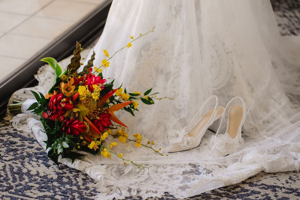 Bridal bouquet of red and yellow flowers beside white heels on lace wedding dress train.