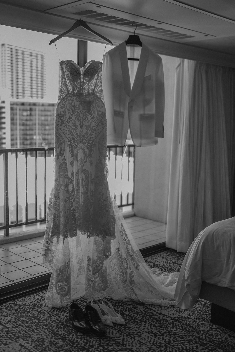 Black and white photo of lace wedding dress and white tuxedo jacket hanging on hotel balcony.