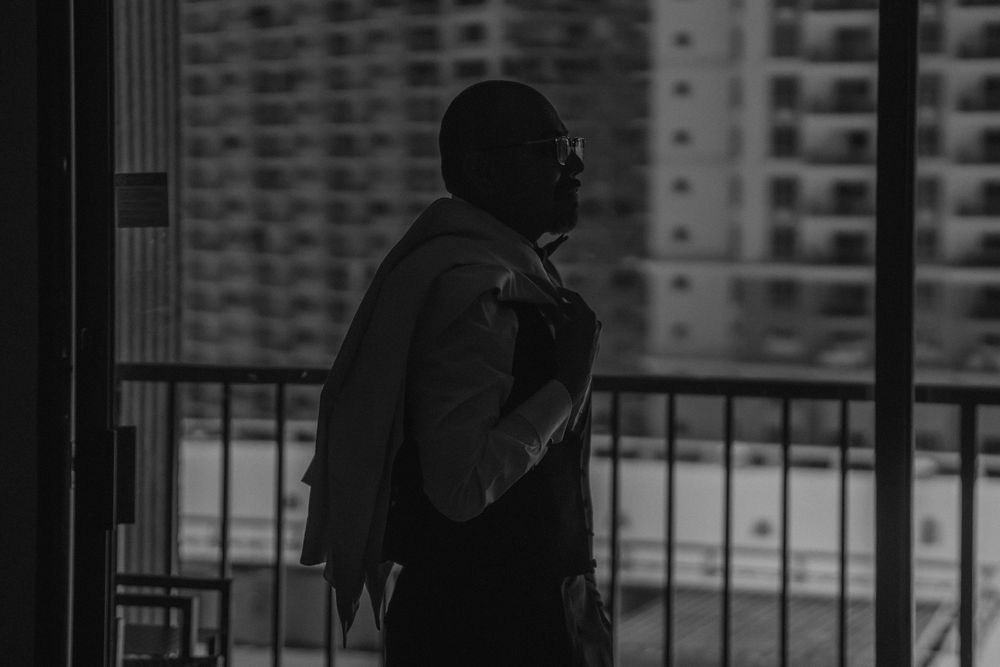 Black and white photo of a man in a suit standing on a balcony with city buildings in the background.