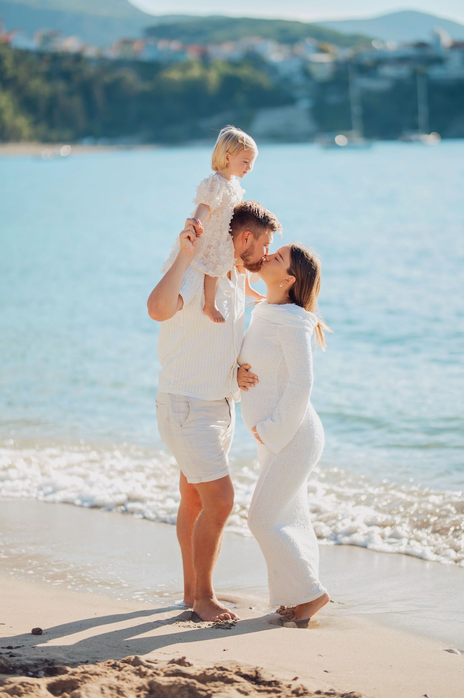 Happy family in white outfits kissing on a sunny beach, father lifting toddler, with turquoise sea in background.