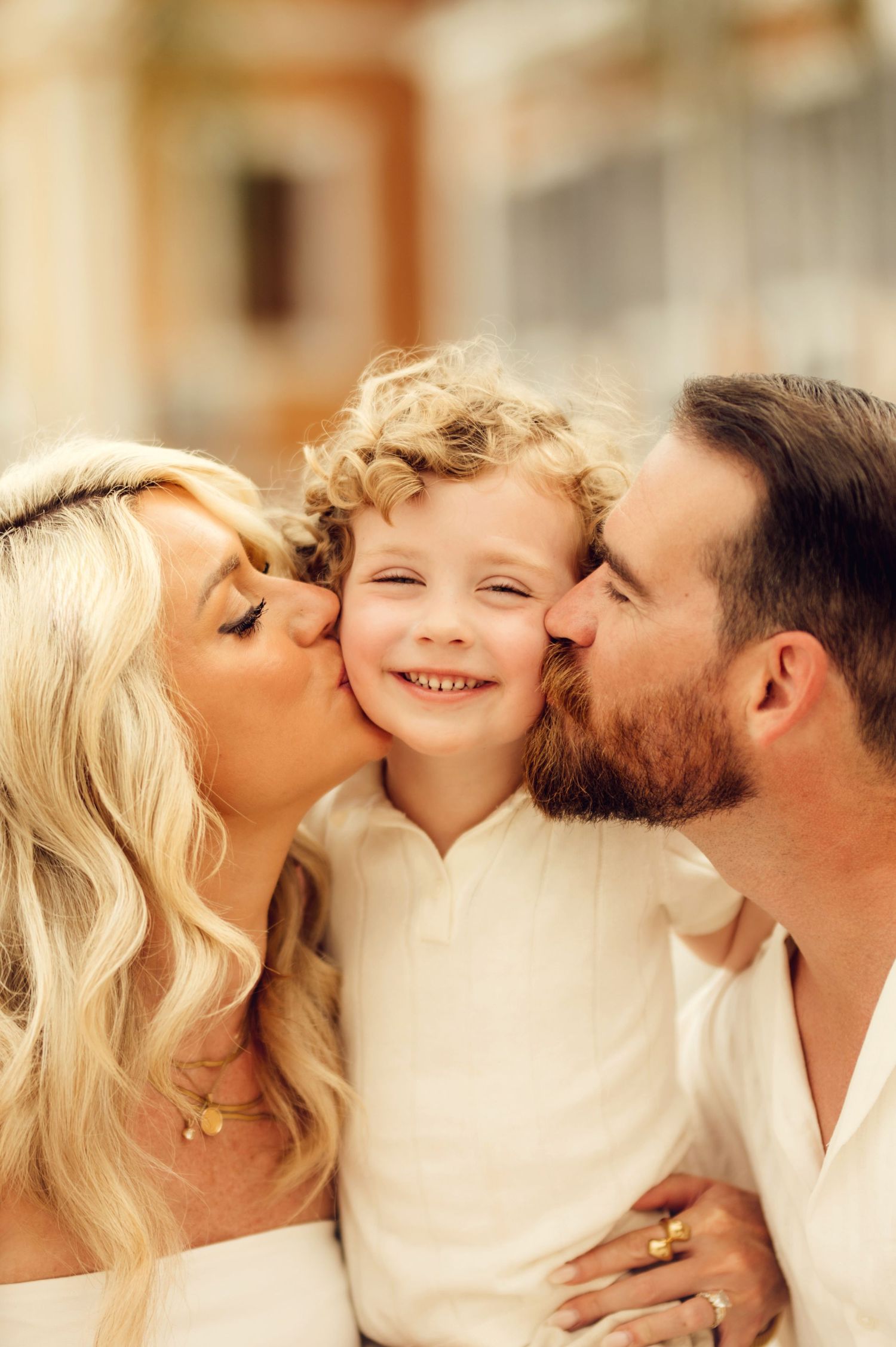 Parents kissing their curly-haired toddler on both cheeks while child smiles joyfully in matching white outfits.