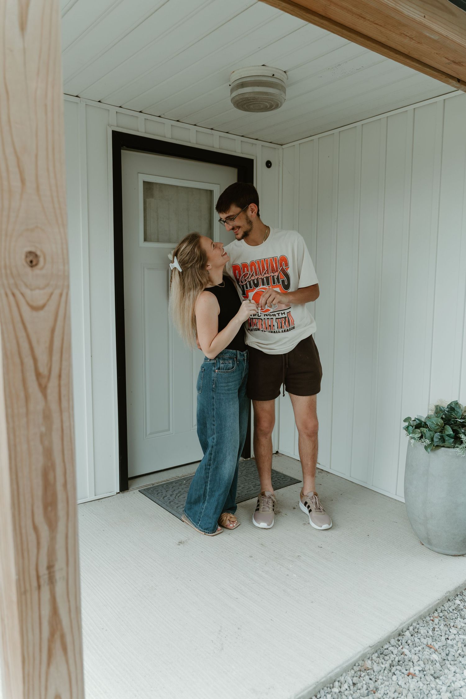 A couple shares a romantic moment on a white porch, woman in jeans and man in graphic tee, standing by a black door.