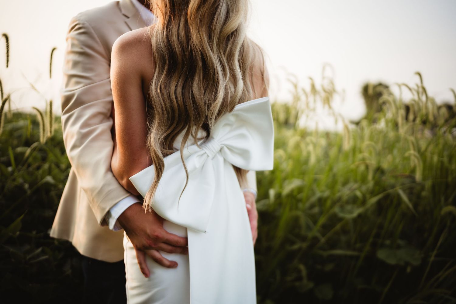 Couple embracing in a field, bride wearing a white gown with a large bow, groom in a tan suit at sunset.