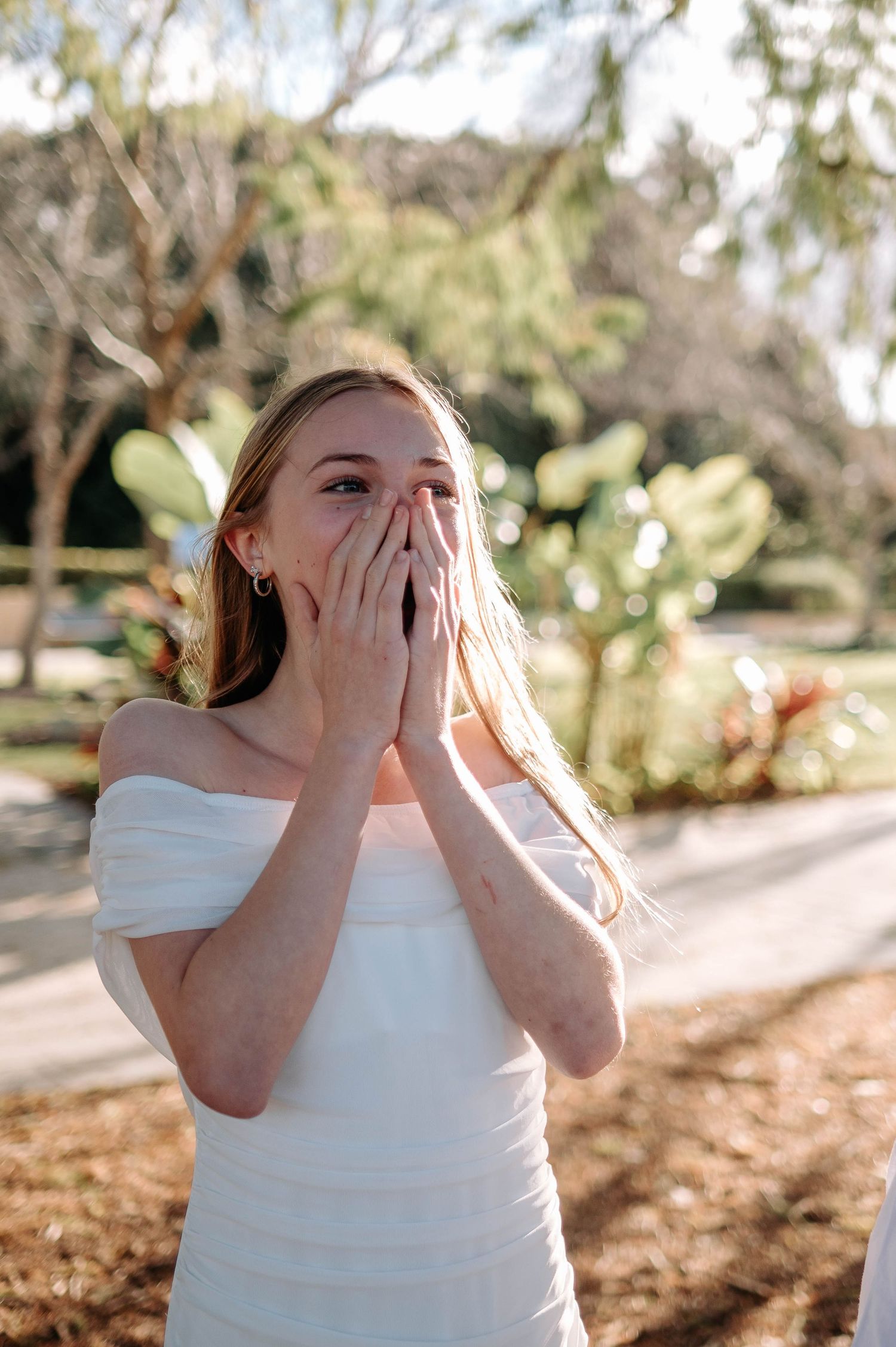 A surprised young woman in a white off-shoulder dress covers her mouth with both hands outdoors near trees.