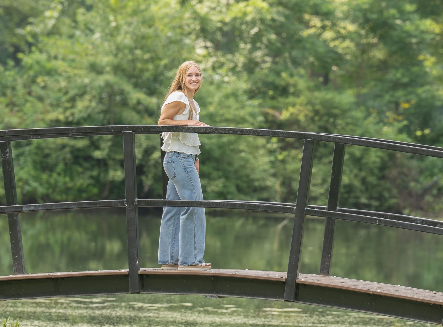 Woman in jeans smiling on a wooden arched bridge over a pond surrounded by lush green trees.