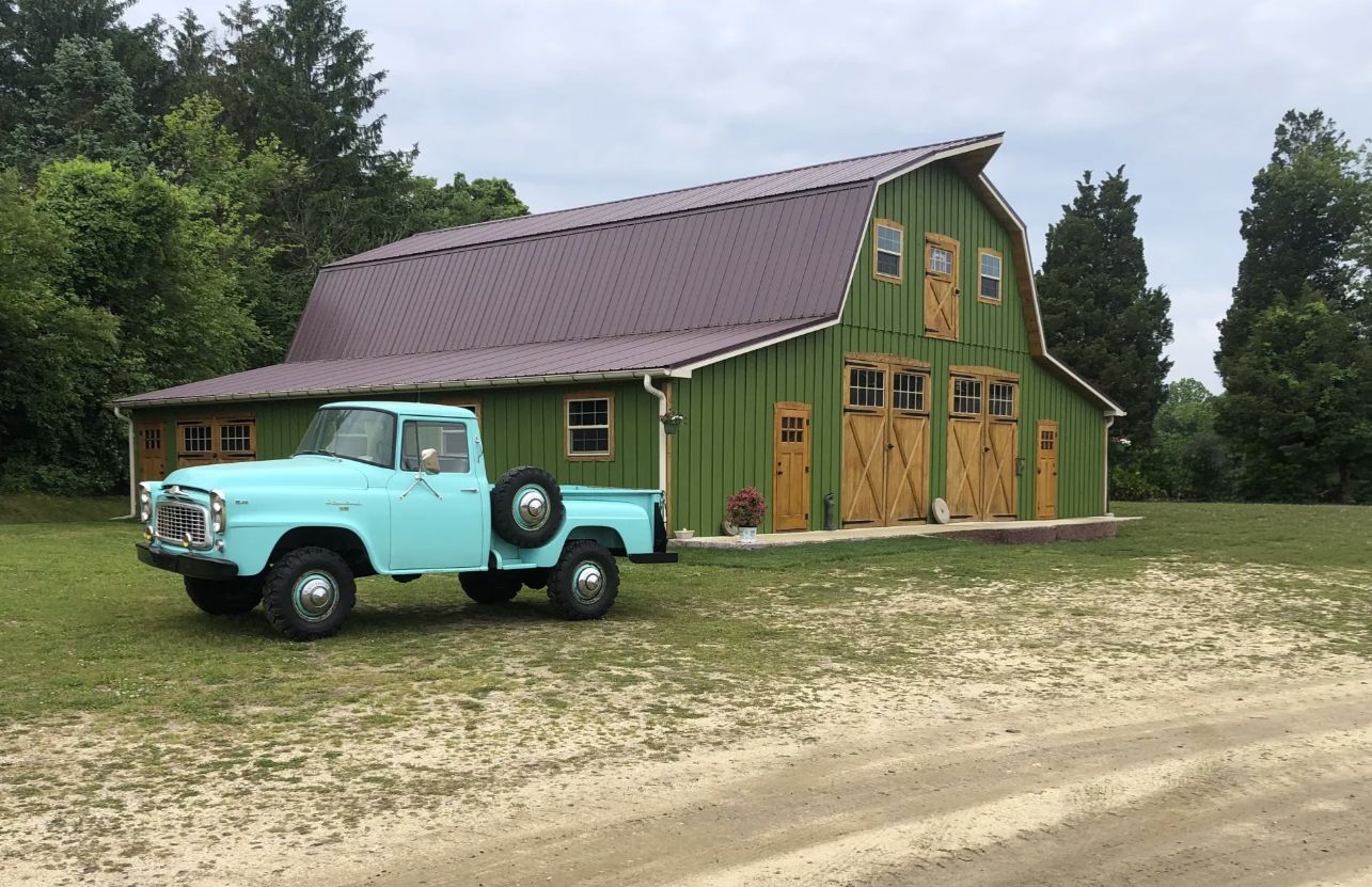 Vintage turquoise pickup truck parked in front of a large green gambrel barn with wooden doors on a gravel driveway.