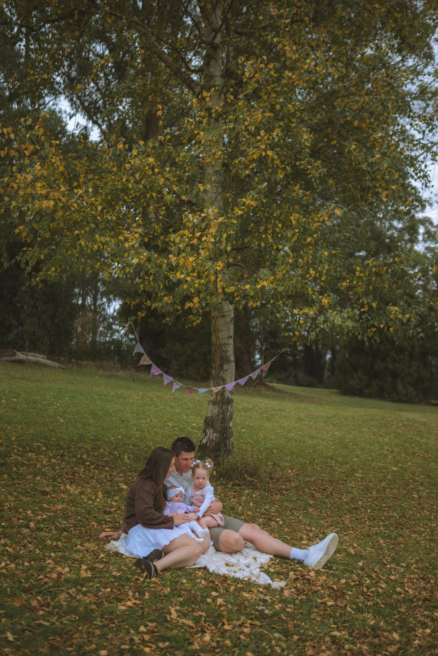 Family sitting on blanket under autumn tree with bunting, enjoying a peaceful outdoor picnic together.