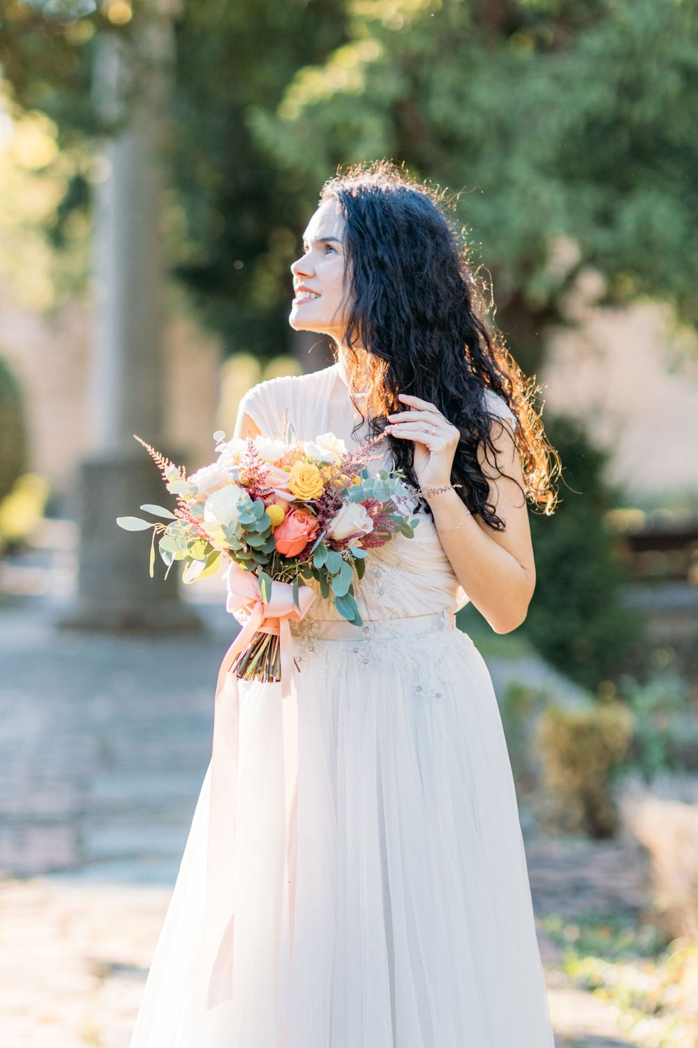 Smiling bride with dark curly hair holding a colorful bouquet in a white gown outdoors.