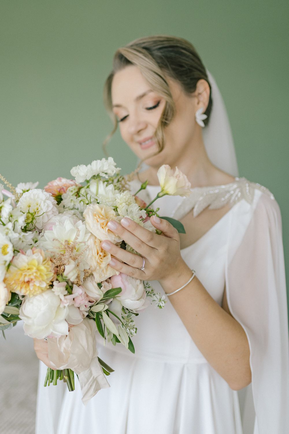 Bride in white gown holding lush pastel bouquet of peonies and roses, smiling softly against sage green background.