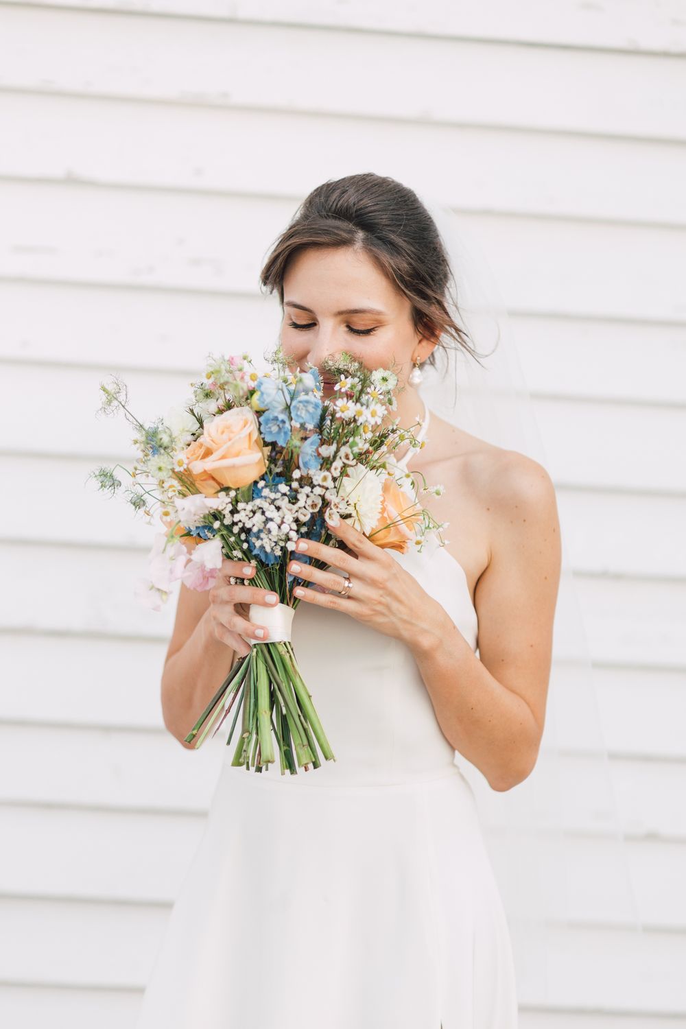 Bride in white strapless gown smelling a pastel bouquet of peach roses and blue flowers against a white backdrop.