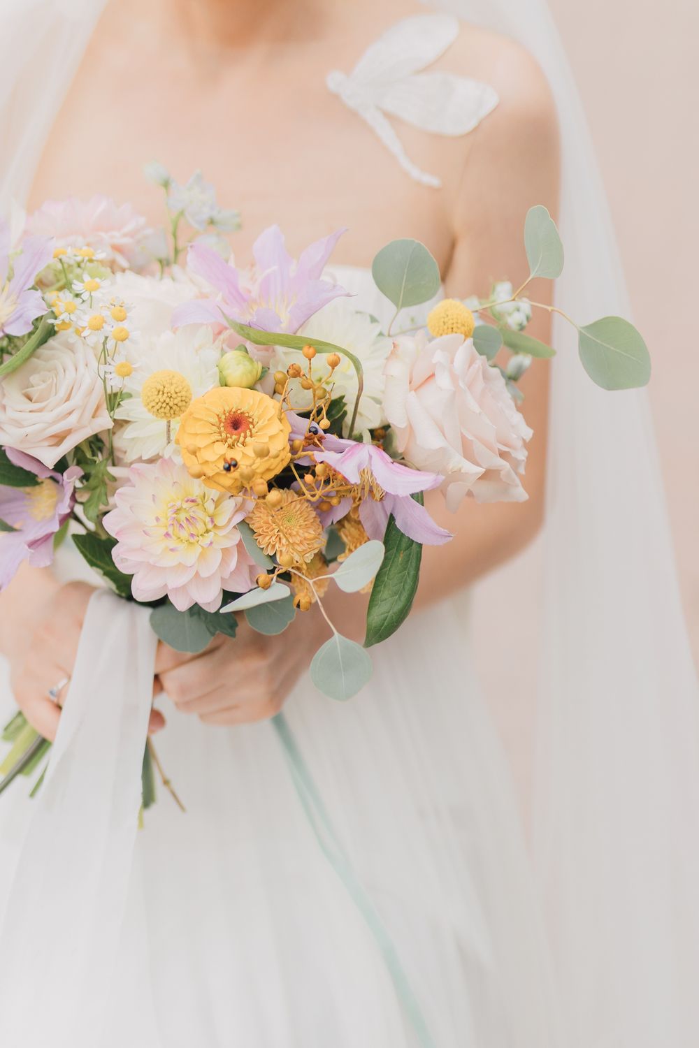 Bride holding a pastel wedding bouquet with yellow dahlias, pink roses, and lavender blooms in a white gown.