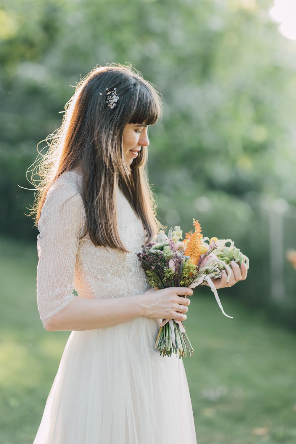 Smiling bride in white lace gown holding colorful wildflower bouquet outdoors in sunlit garden.